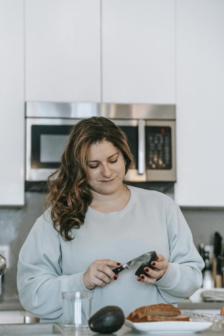 Optimistic female cook cutting ripe avocado while making toasts in modern light kitchen with microwave oven at home during breakfast